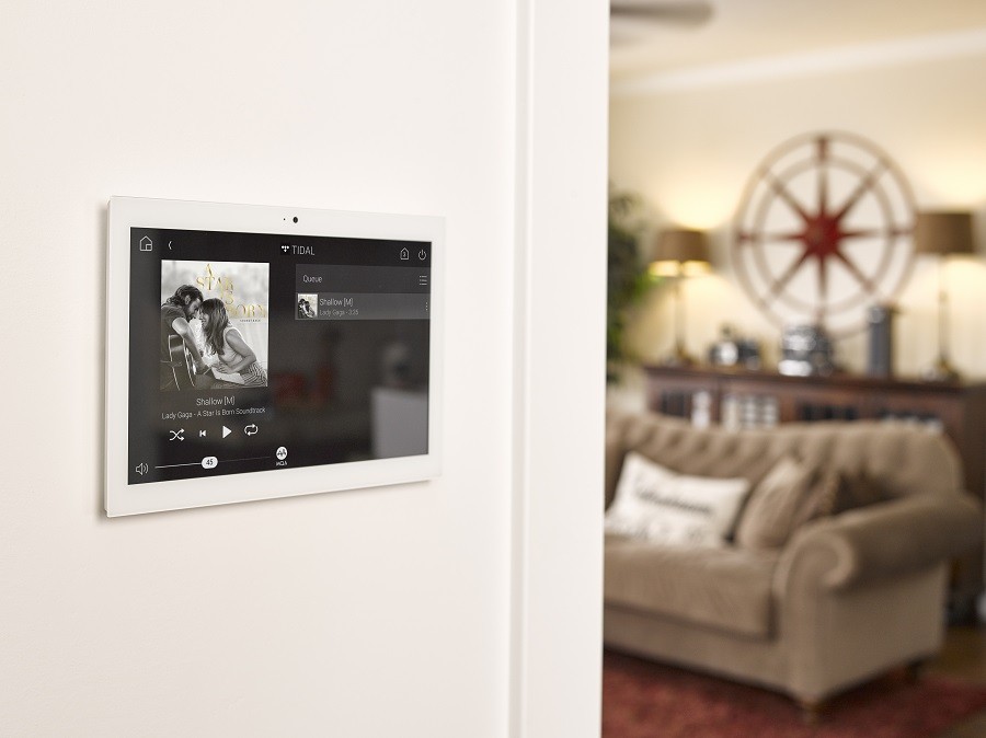 A wall-mounted Control4 touch screen displaying song information and cover art with the living room in the background. 