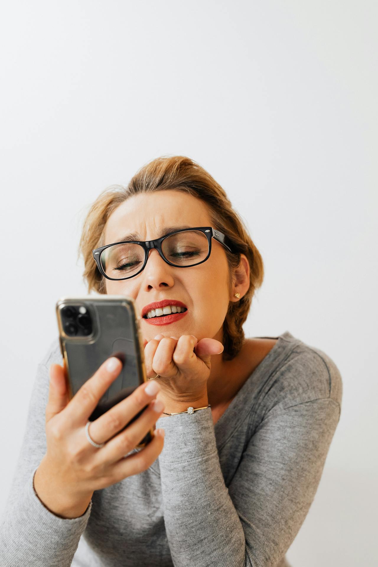 Person wearing glasses and a grey long‑sleeve shirt looking closely at a smartphone against a plain white background.