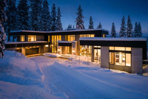 Modern house lighting at night surrounded by snow and pine trees side view